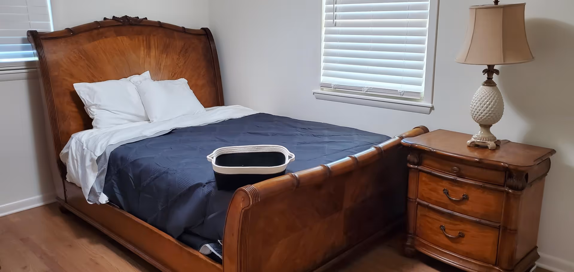 Wooden bed with navy comforter, nightstand lamp, and laundry basket in a bright bedroom.