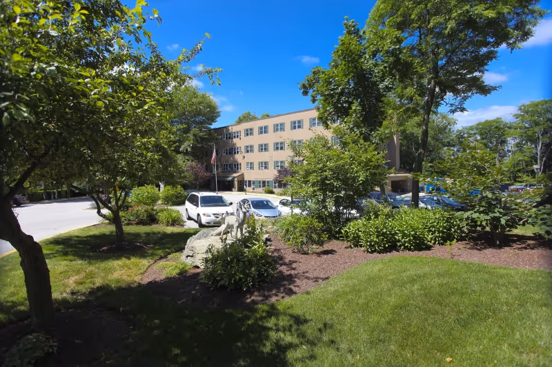 Front exterior of a multi-story rehabilitation and healthcare building with landscaped grounds, trees, and parked cars.