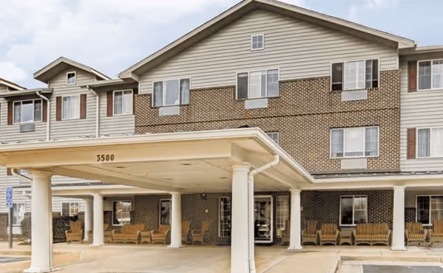 Front exterior view of a three-story senior living facility building with a covered entrance supported by white columns. Several wooden rocking chairs are placed under the covered area and along the front porch. The building has a combination of brick and siding exterior with multiple windows.