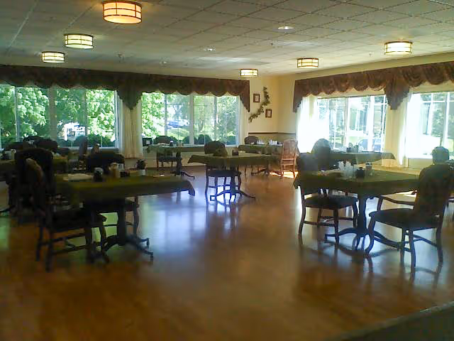 A spacious dining room with several tables and chairs arranged throughout. Large windows with curtains allow natural light to fill the room, and the floor is made of polished wood. The room has ceiling lights and a calm, inviting atmosphere.