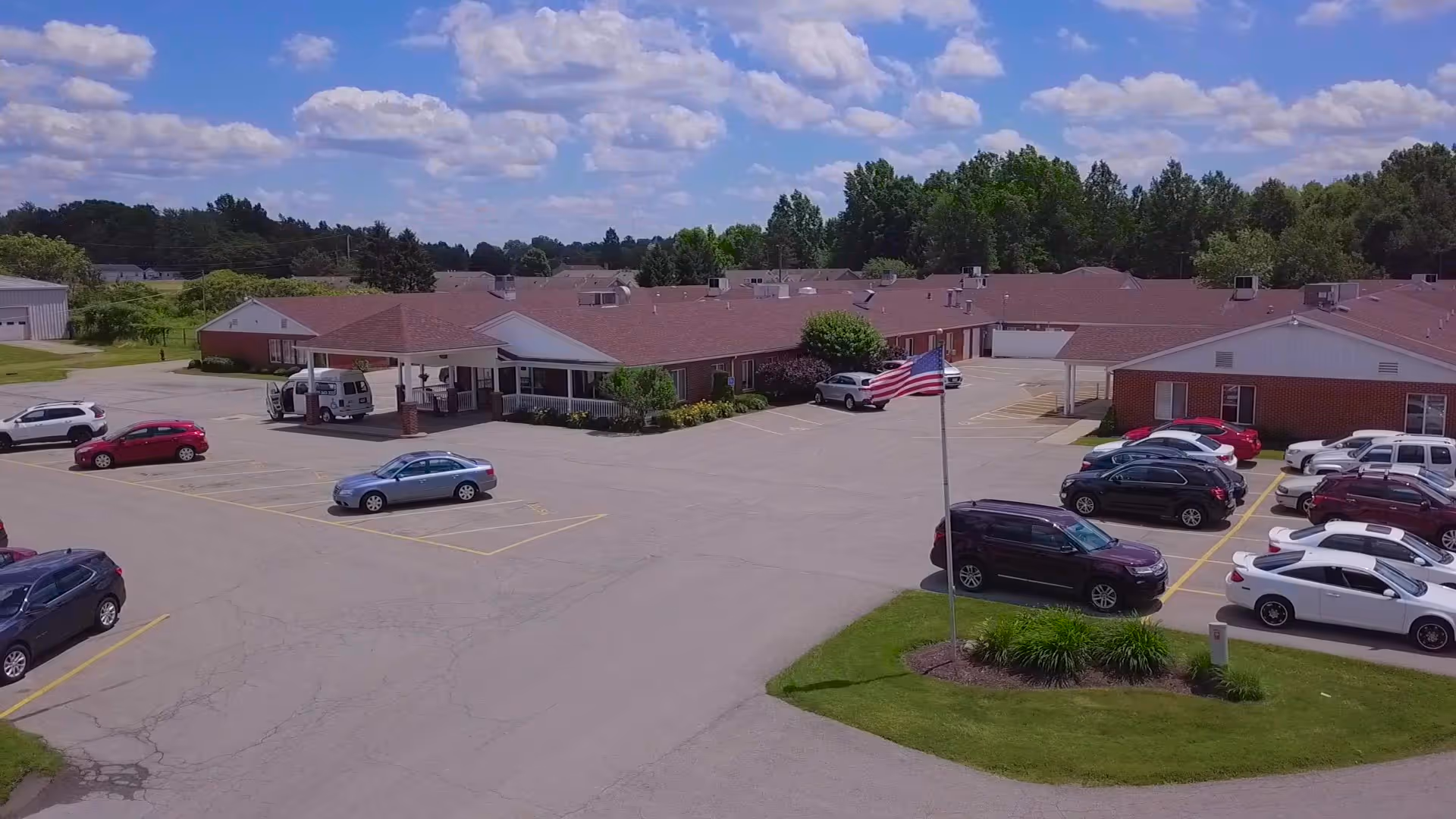 Aerial view of a single-story senior living facility with a large parking lot in front. Several cars are parked, and an American flag is flying on a flagpole near the entrance. The building has a red roof and brick exterior, surrounded by trees and greenery under a partly cloudy sky.