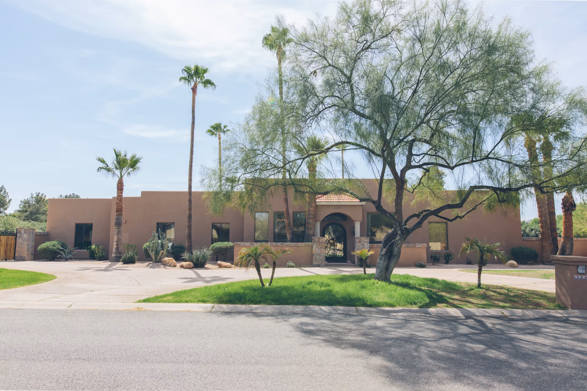 Front exterior view of a single-story building with a southwestern architectural style, featuring a tiled roof entrance, several windows, and surrounded by palm trees and desert landscaping under a clear sky.