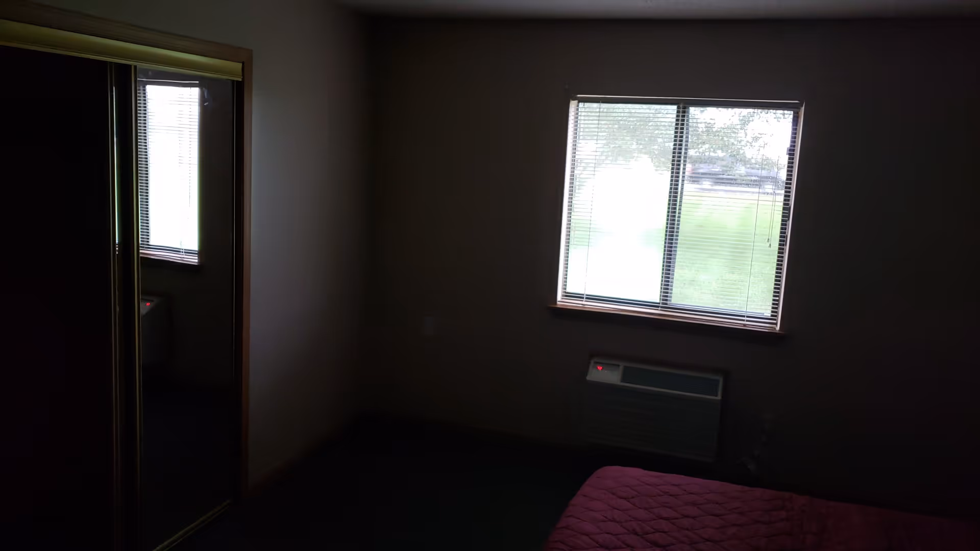 Dim bedroom with pink quilt on bed, window blinds, and wall-mounted heater.