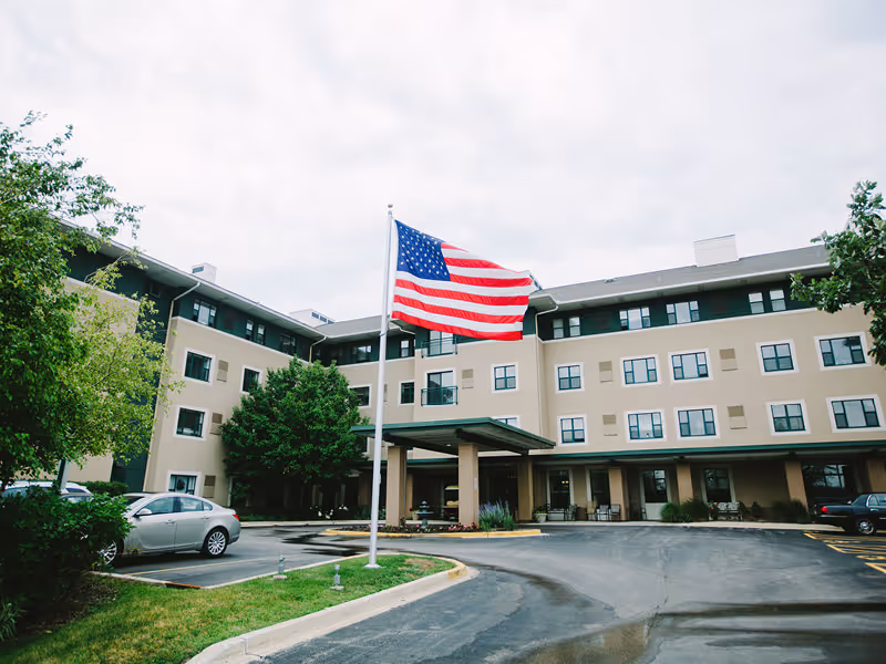Exterior view of a three-story senior living facility building with multiple windows and a covered entrance. An American flag is flying on a flagpole in front of the building, and several cars are parked in the driveway and parking area. Trees and greenery surround the building.
