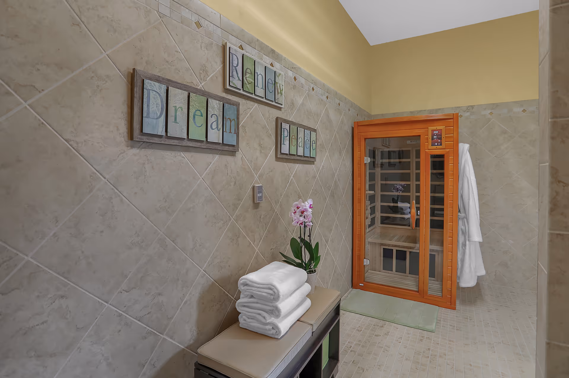 A tiled room with beige walls featuring three framed words: Dream, Renew, and Peace. There is a small bench with folded white towels and a potted orchid on top. To the right, there is a wooden infrared sauna with a glass door and a white robe hanging on the wall next to it.
