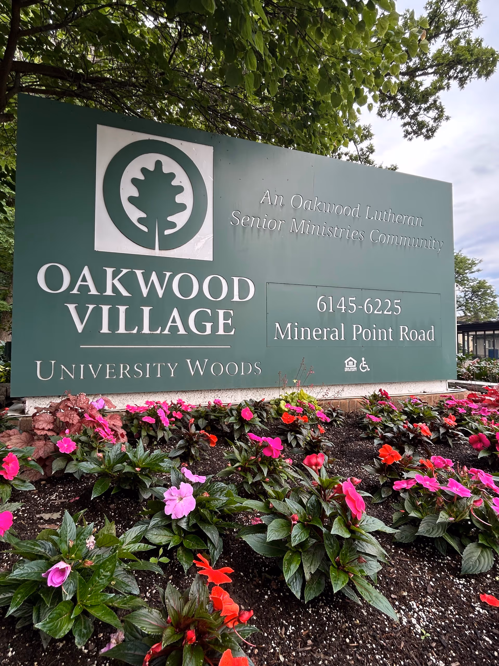 Large green Oakwood Village sign with colorful flowers along a garden bed in front.