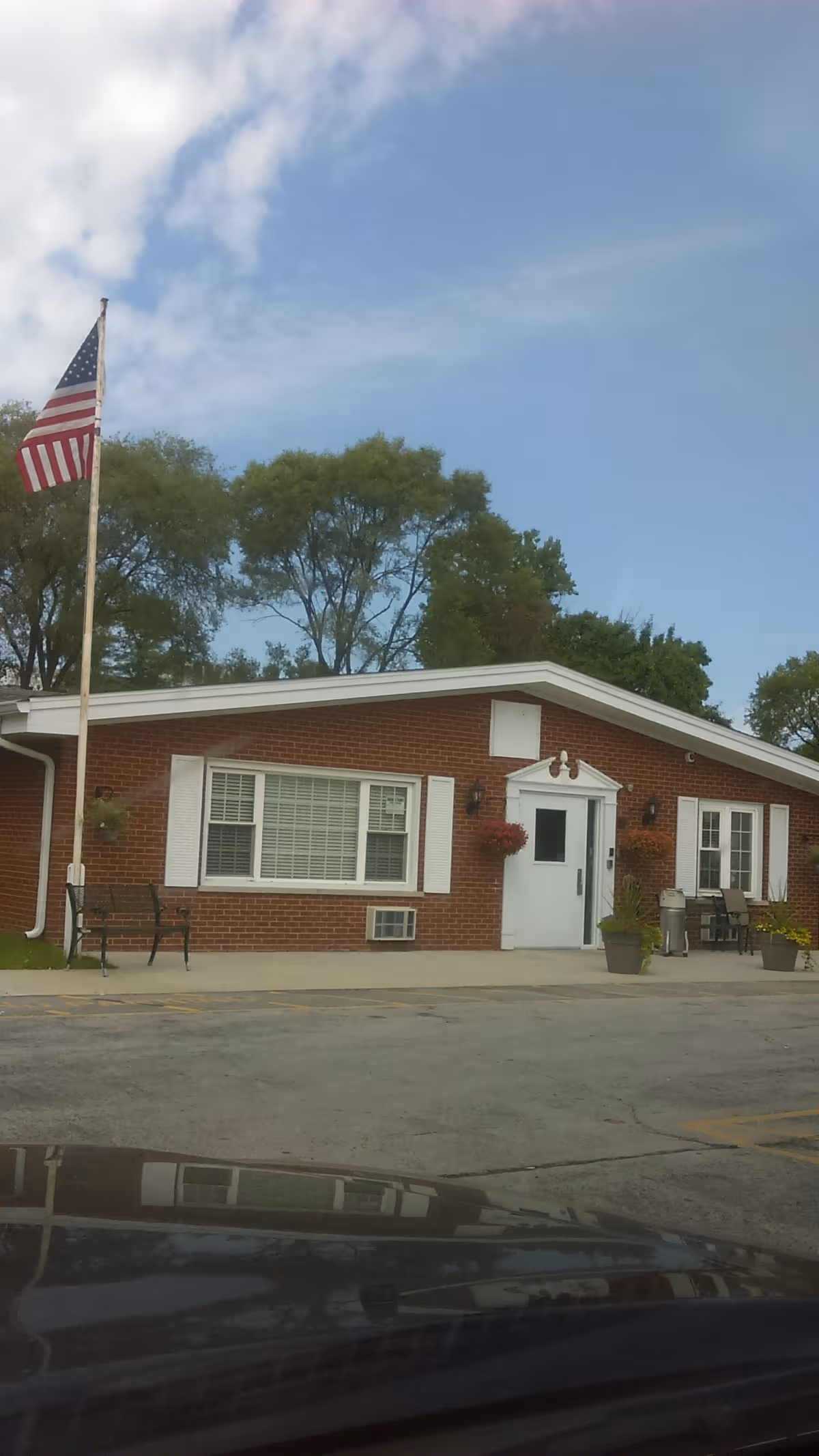 Single-story brick senior living facility entrance with white door, windows, seating, and American flag.