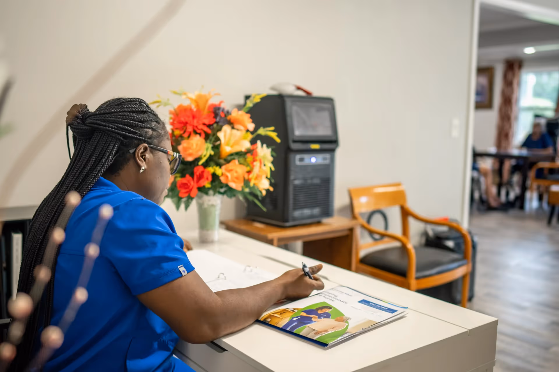 A woman in a blue uniform is sitting at a white desk, writing in a booklet. Behind her, there is a vase with colorful flowers and a wooden chair. In the background, there is a room with more chairs and a person sitting at a table.