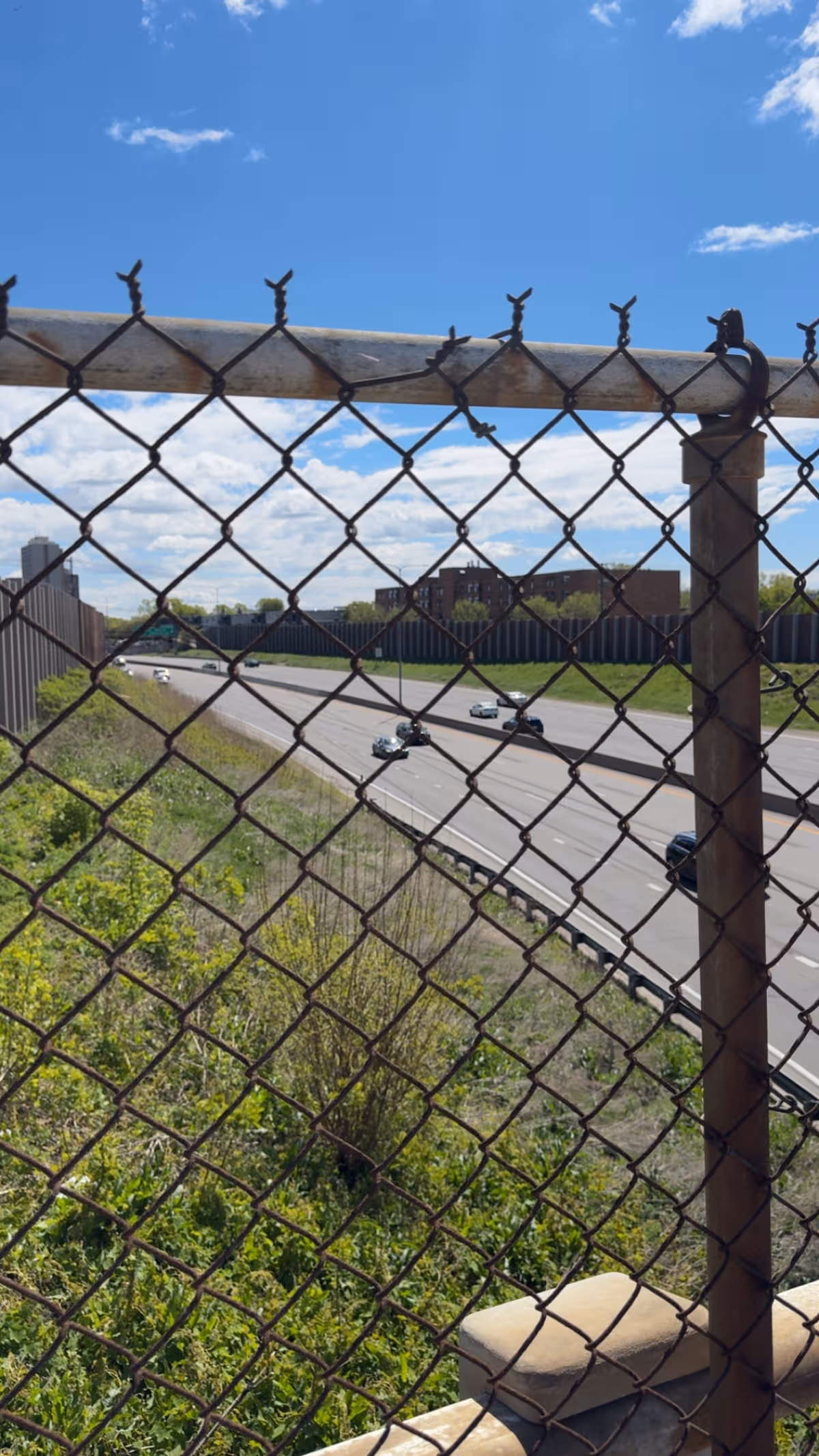 View through a rusty chain-link fence over a highway and green area.