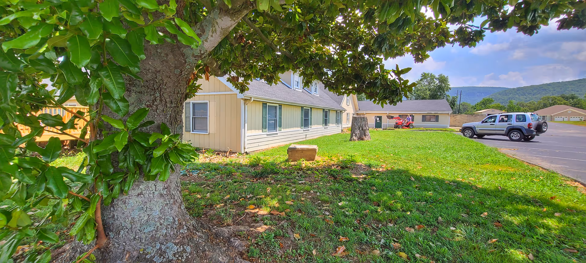 Senior living facility exterior with beige building, large tree, lawn, and parked vehicles.