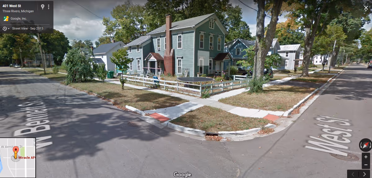 Residential street corner with a small fenced yard and pastel houses seen from street view.