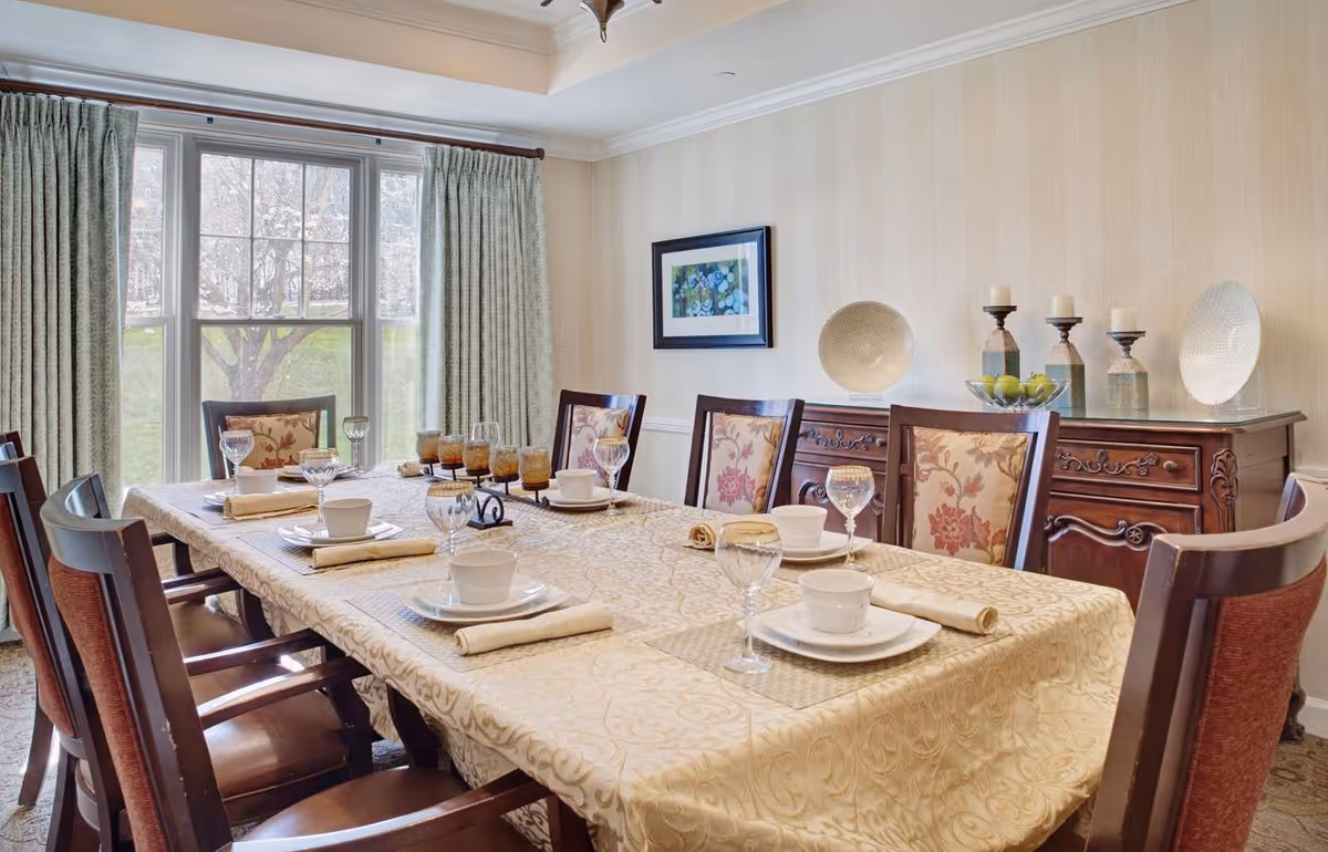 Dining room with a long set table, upholstered chairs, a large window, and a decorative sideboard.