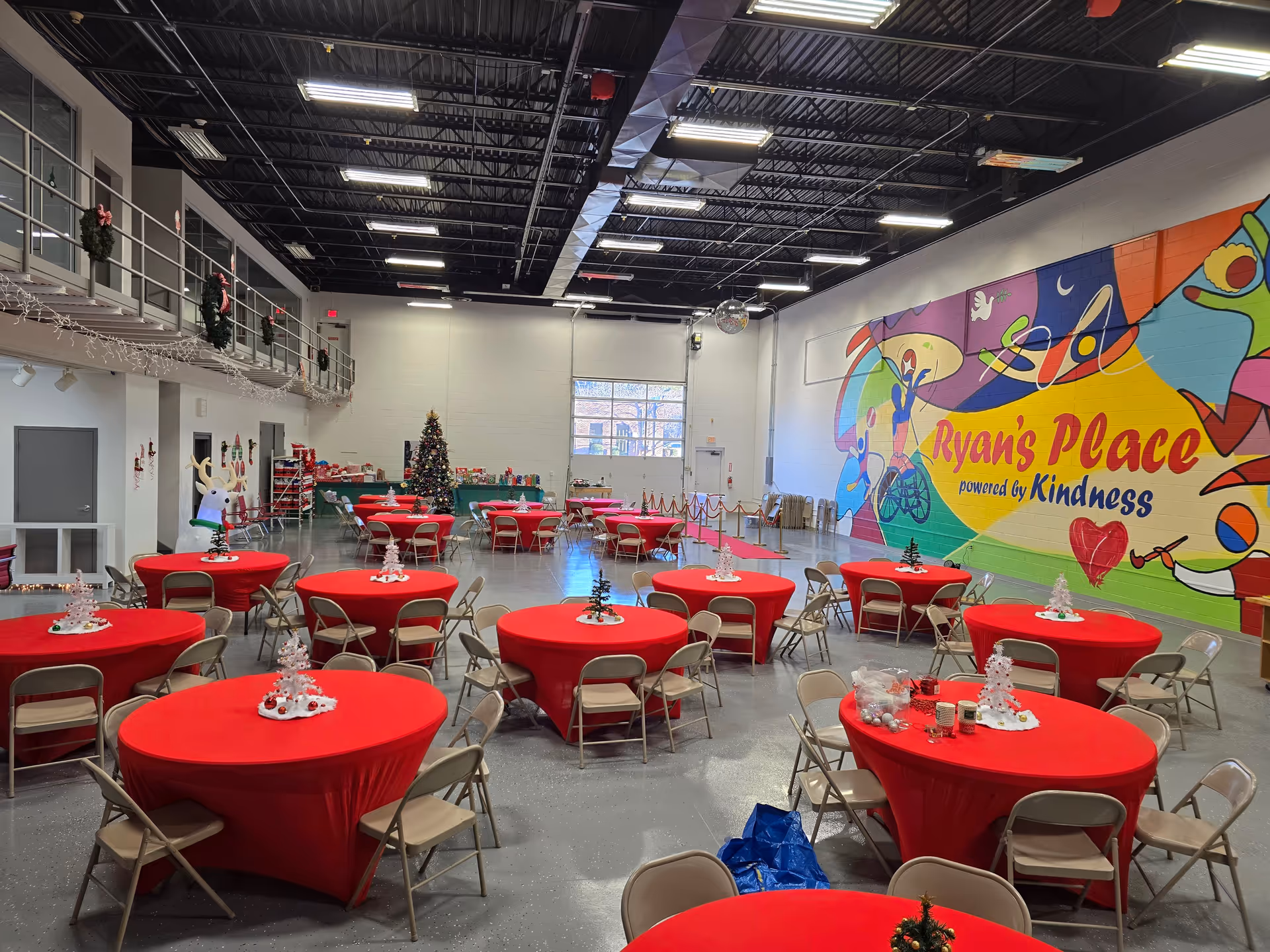 Spacious indoor hall with red-covered round tables, chairs, and a colorful mural reading Ryan's Place.