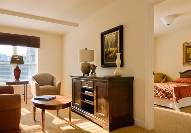 A cozy living room area with a round wooden coffee table, an armchair, a side table with a lamp, and a wooden cabinet with decorative items and a lamp on top. Sunlight streams through a window with blinds. An adjoining bedroom with a bed covered in a rust-colored quilt is visible through an open doorway.