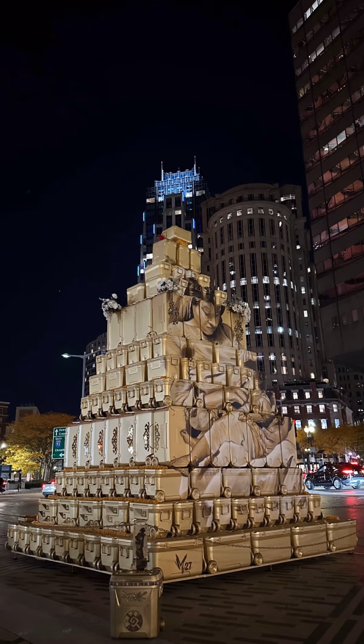 Nighttime city street with a large stacked sculpture made of suitcases.