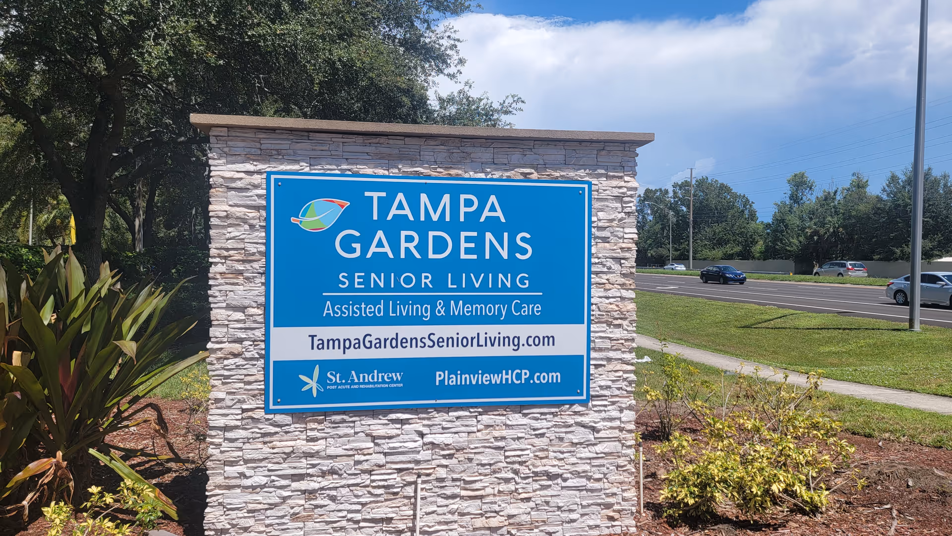 Blue sign for Tampa Gardens Senior Living mounted on a stone pedestal outdoors.