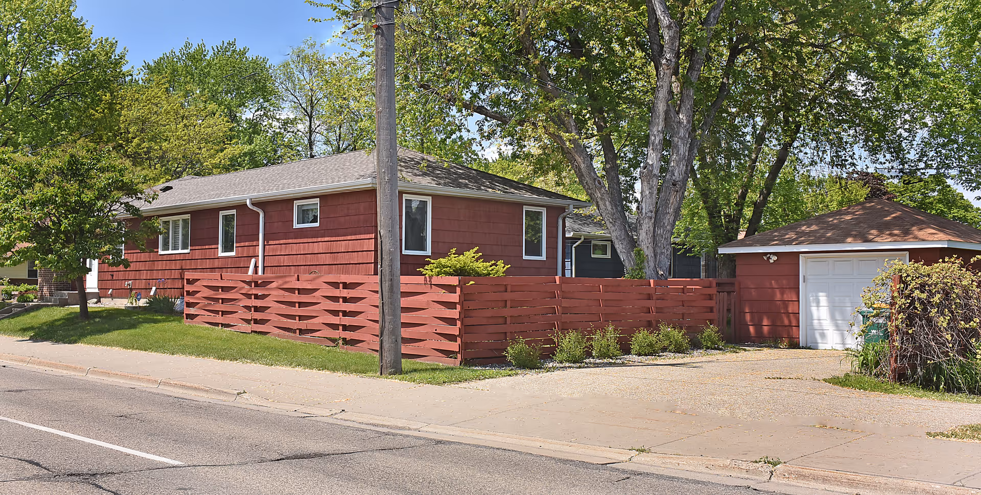 Exterior view of a single-story red building with a matching red wooden fence and a detached garage. The building is surrounded by green trees and grass, with a sidewalk and street in the foreground under a blue sky with some clouds.