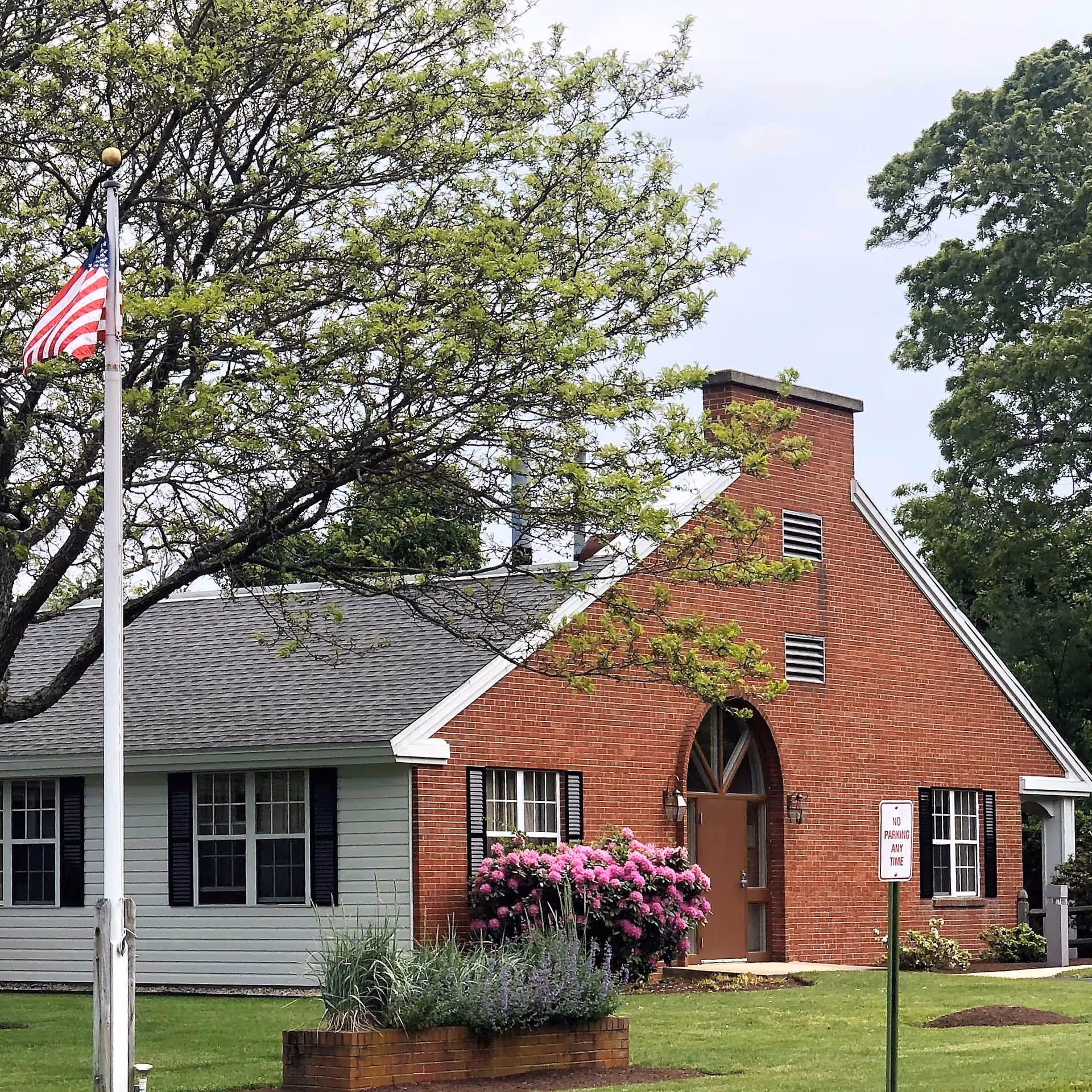 Exterior view of a single-story building with a brick facade and gray siding, surrounded by green grass, trees, and flowering bushes. An American flag is flying on a flagpole in front of the building, and a 'No Parking Any Time' sign is visible near the entrance.