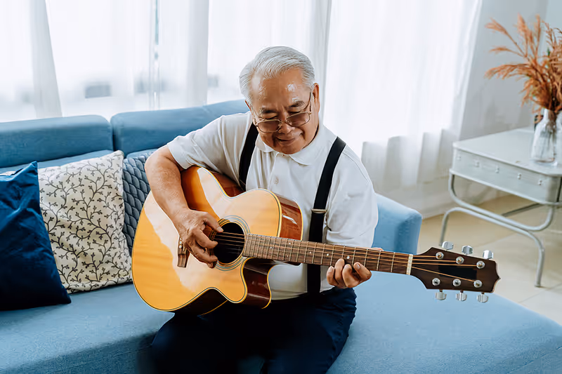 An elderly man with gray hair and glasses is sitting on a blue couch playing an acoustic guitar. The room is bright with natural light coming through sheer white curtains. There are decorative pillows on the couch and a small white table with a vase of dried flowers in the background.