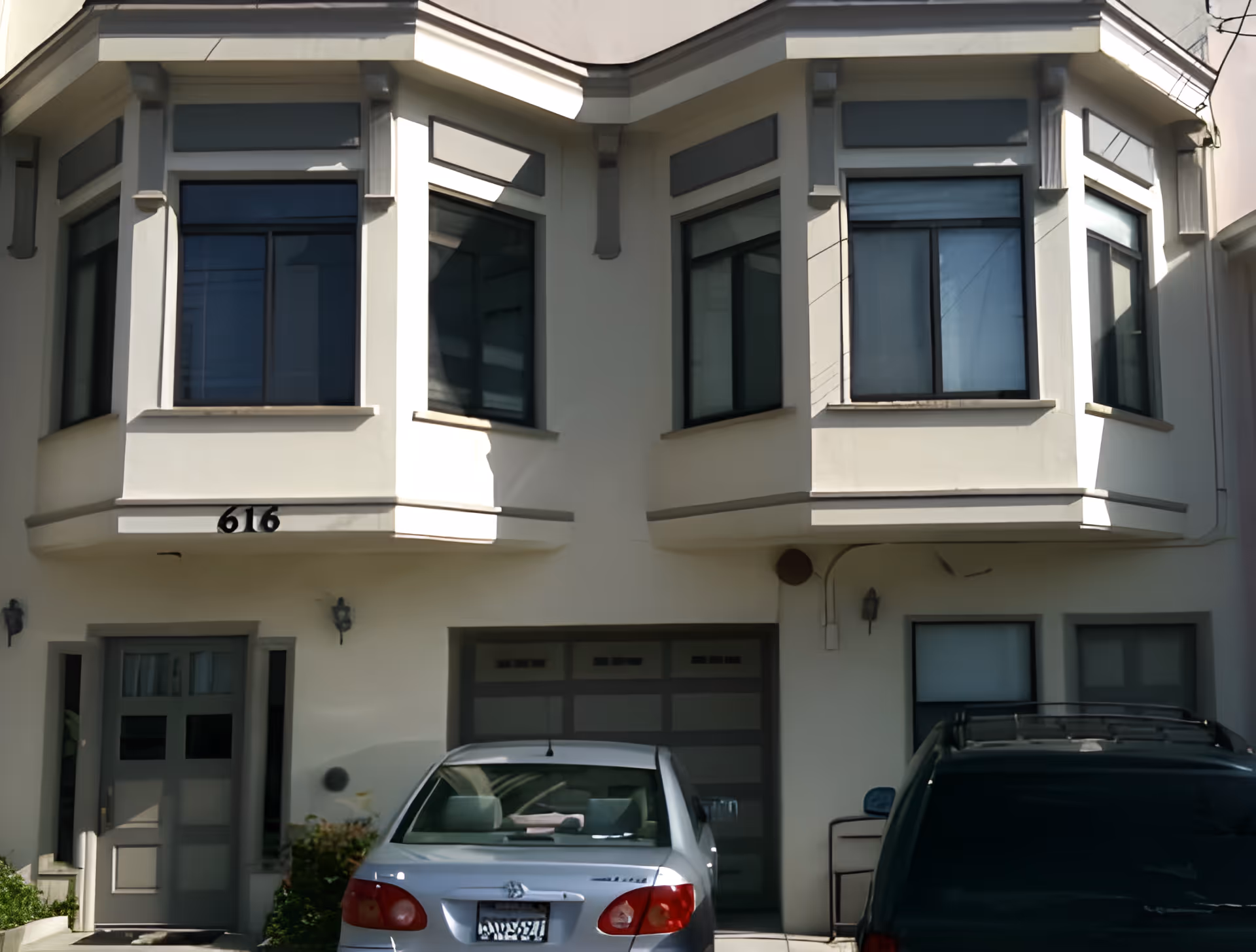 Front facade of a light-colored two-story building with bay windows, a garage and two parked cars.