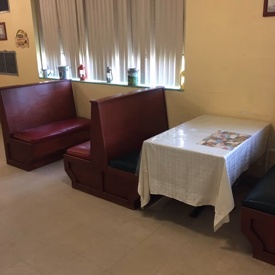 A dining area with two wooden booth-style benches with red cushions facing each other across a table covered with a white tablecloth. The table has a colorful placemat on it. Behind the benches is a window with vertical blinds and several decorative lanterns and containers on the windowsill. The walls are painted yellow.