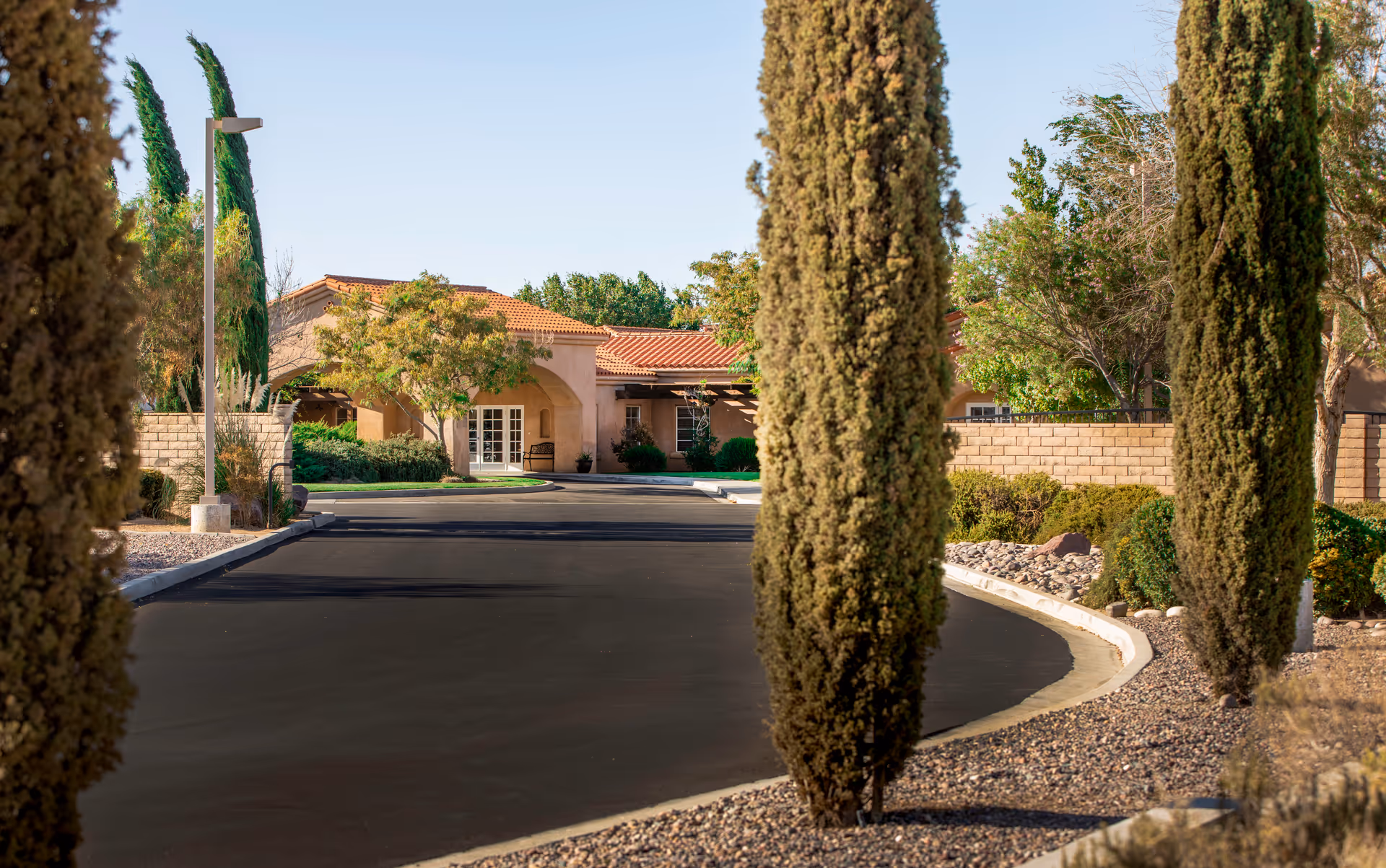 Curved driveway leads to a beige tile-roofed building, framed by tall trees and desert landscaping.