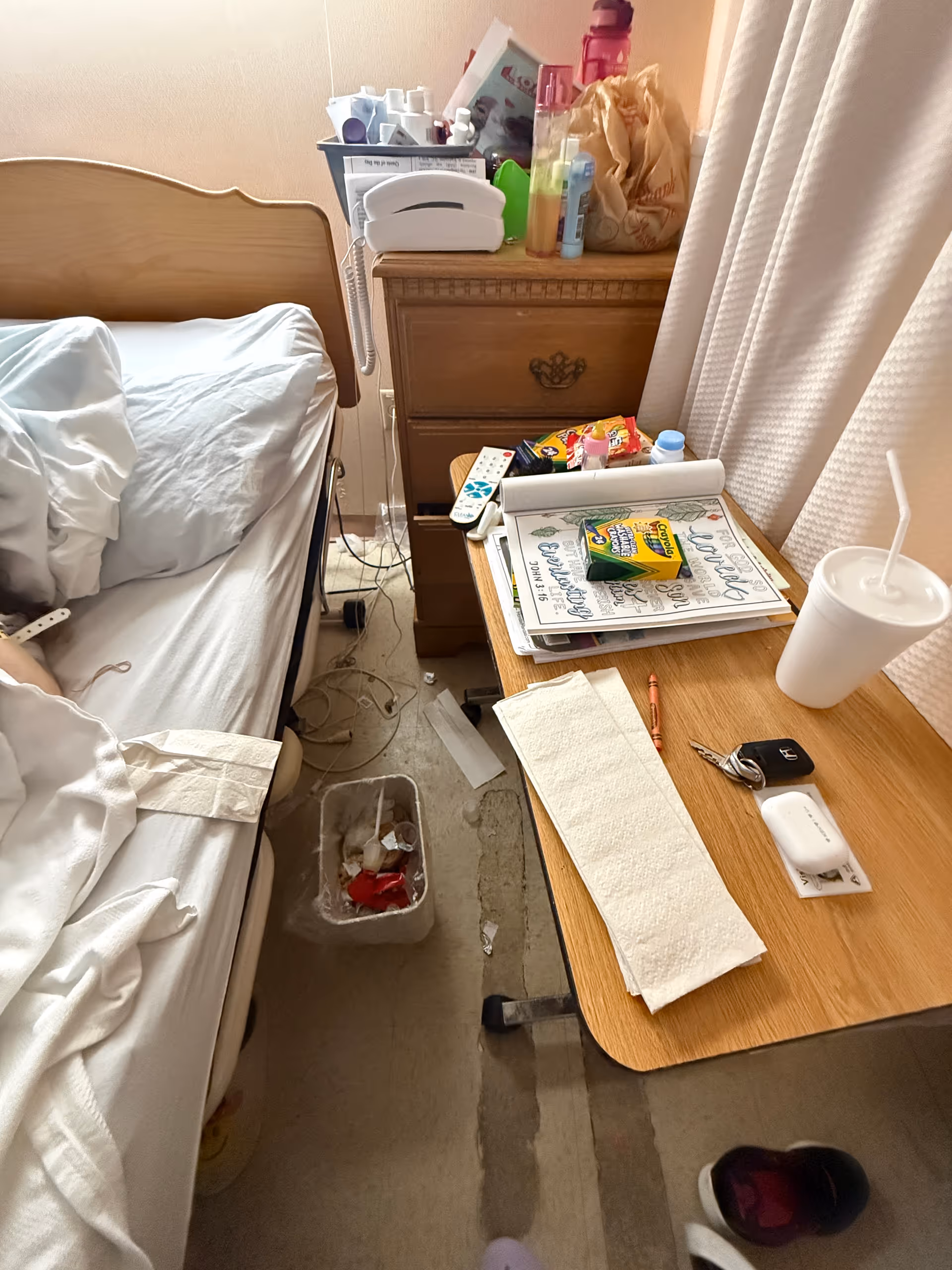 A cluttered hospital-style bedroom with a bed, nightstand, papers, and personal items.