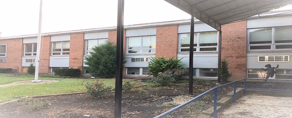 Exterior view of a single-story brick building with multiple windows, a small garden area with bushes and plants, a flagpole, and a covered walkway with metal support beams.