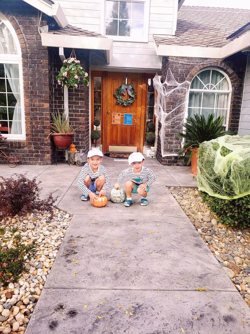Two young children crouch on the front walkway holding pumpkins outside a brick house entrance decorated with a wreath and Halloween cobwebs.