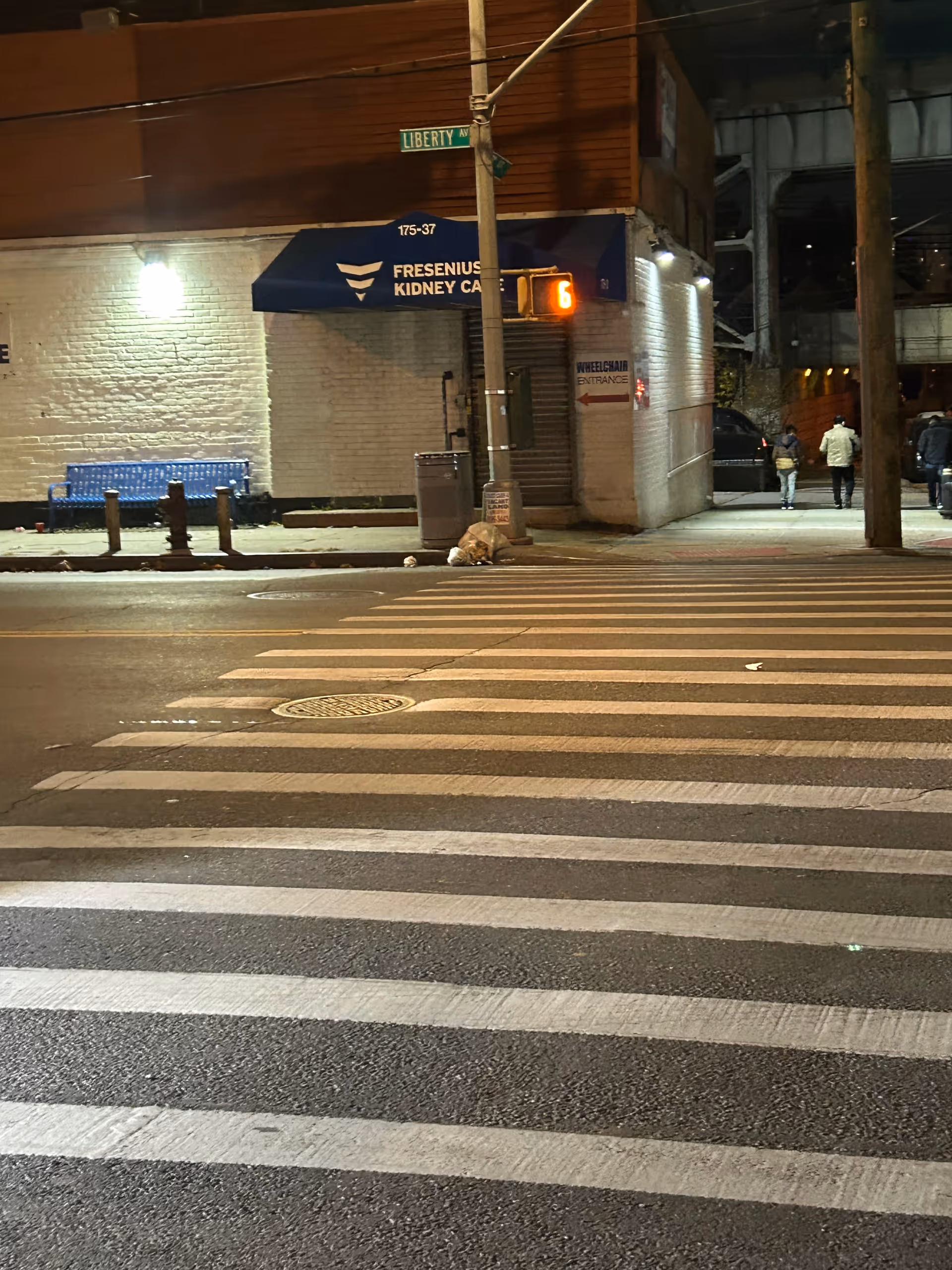 Night street scene with a white brick building, blue awning, crosswalk, and pedestrians.