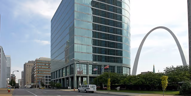 Tall glass office building beside a large arch, with a city street and trees.