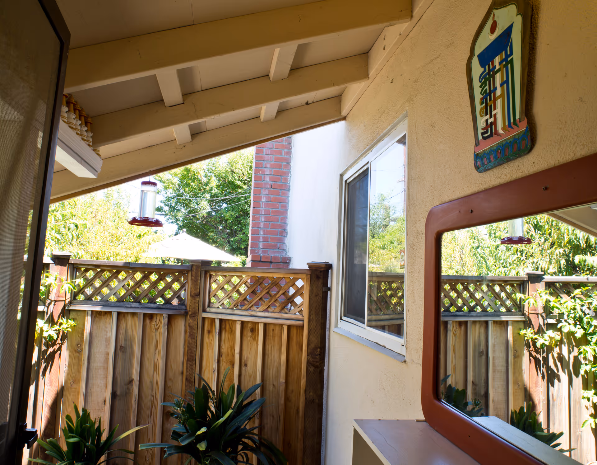 Covered backyard patio with wooden fence, potted plants, bird feeder, window, and decorative wall mirror.
