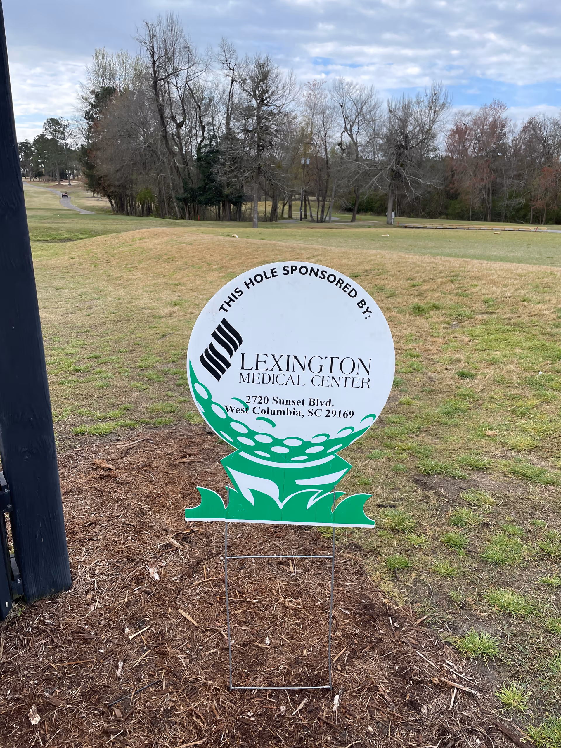 White circular sign on metal stand advertising a hole sponsor on a grassy golf course.