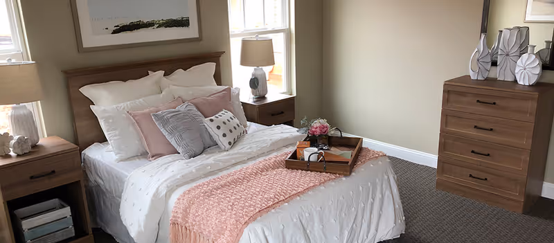 A cozy bedroom with a neatly made bed featuring white and pink bedding, multiple pillows, and a pink throw blanket. There are two wooden nightstands with lamps on either side of the bed, a wooden dresser with decorative vases, and a framed picture hanging above the headboard. The room has beige walls and carpeted flooring.