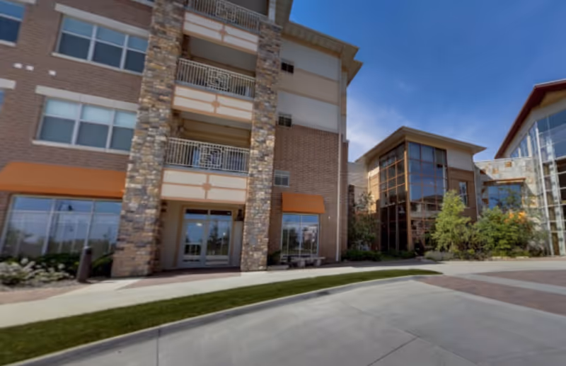 Exterior view of a multi-story senior living facility building with brick and stone facade, large windows, balconies, and a landscaped area with shrubs and trees under a clear blue sky.