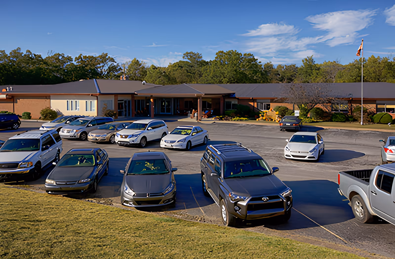 Exterior view of Ahc West Tennessee Transitional Care facility showing a single-story building with a covered entrance, surrounded by a parking lot with several parked cars and a flagpole on the right side. Trees and a blue sky with some clouds are visible in the background.
