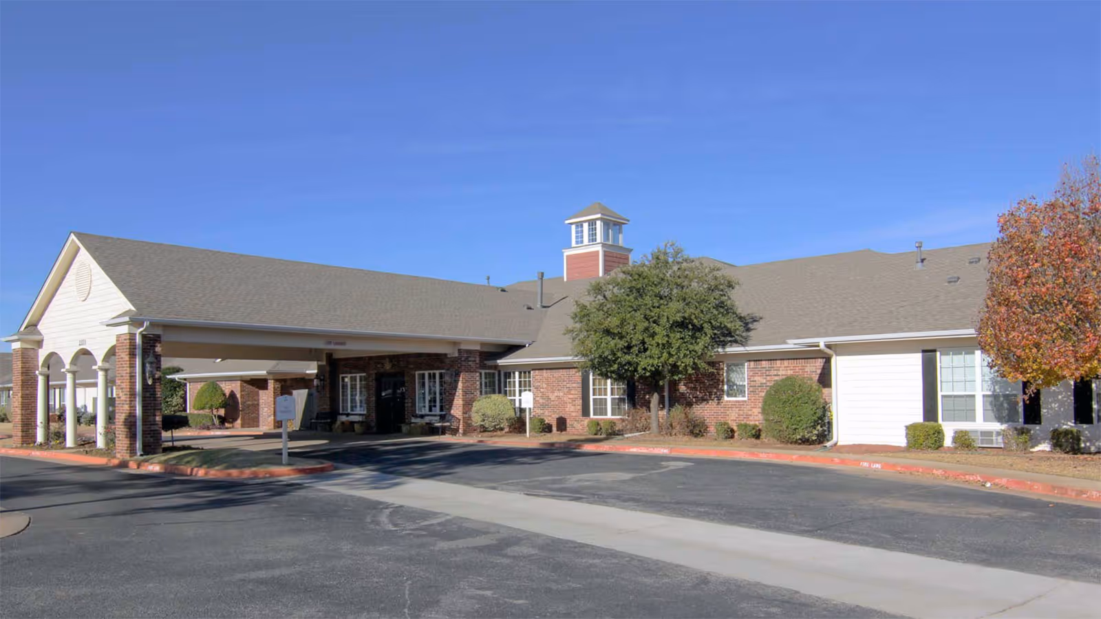 Exterior view of Radiance Senior Living at The Village, showing a single-story brick building with a covered entrance, a small tower feature on the roof, and landscaped bushes and trees under a clear blue sky.
