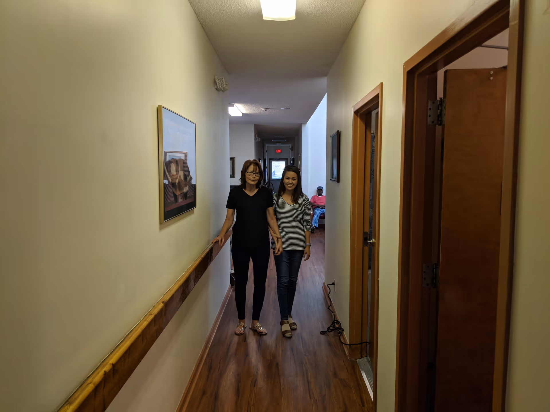 Two women standing and smiling in a hallway with wooden flooring and cream-colored walls. One woman is wearing a black shirt and pants, and the other is wearing a striped shirt and jeans. There is a framed picture on the left wall and an open door on the right. In the background, a person in a pink shirt and blue pants is sitting on a bench.