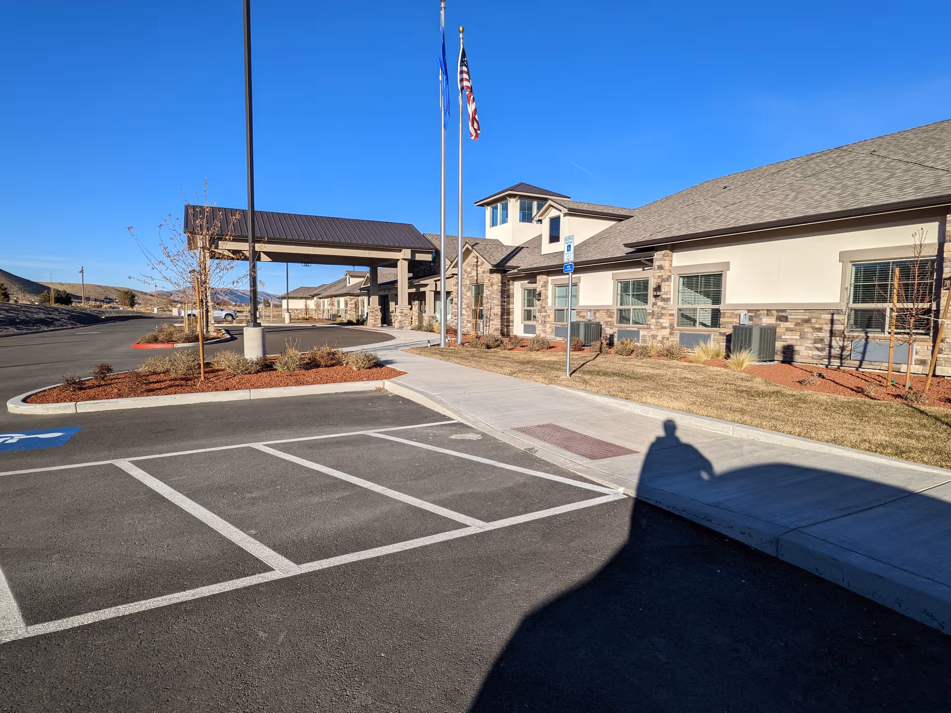 Senior living facility exterior with parking lot, entrance canopy, flagpoles, and clear blue sky.