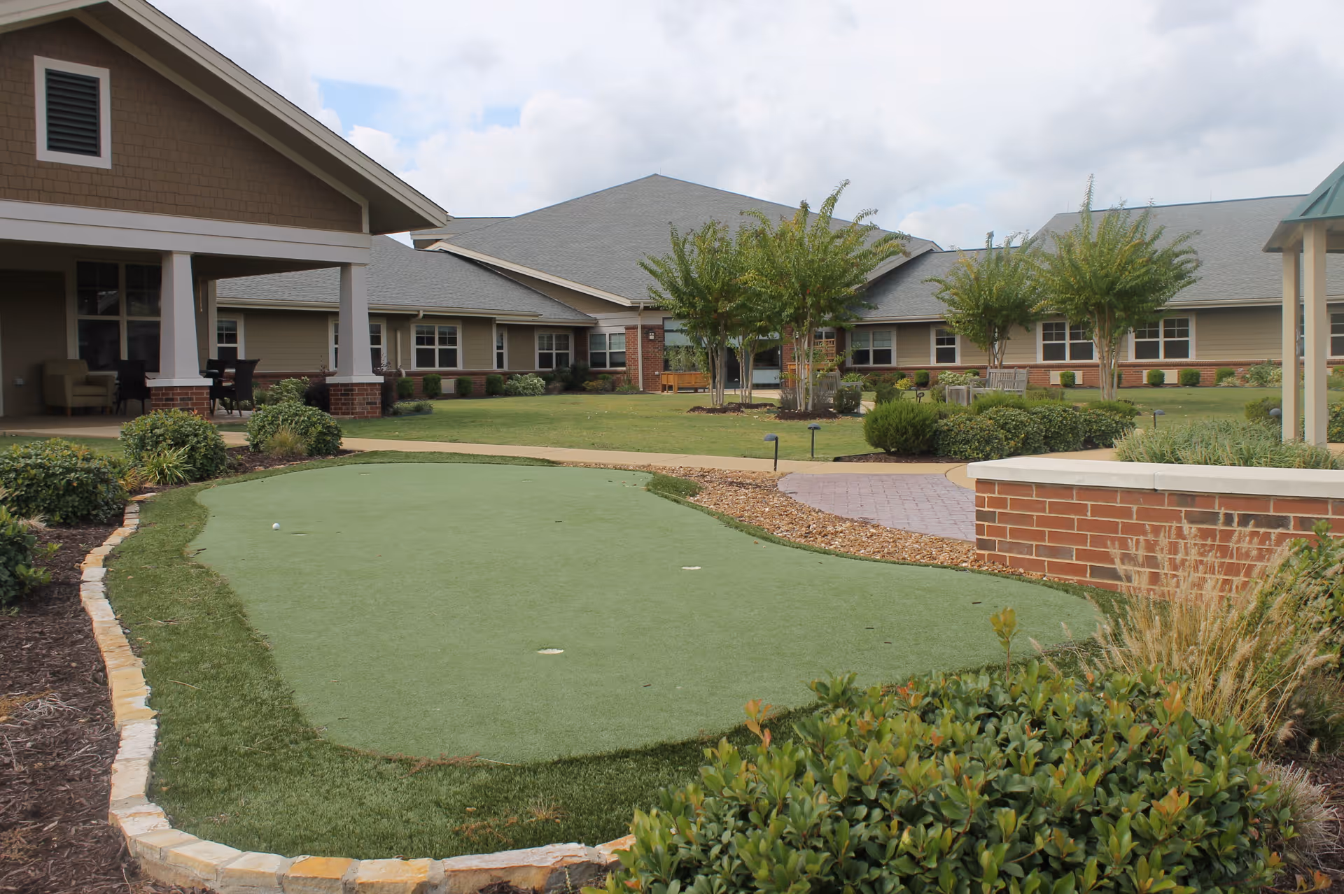 Courtyard with a small putting green, landscaped beds, and a covered patio outside a single-story senior living building.