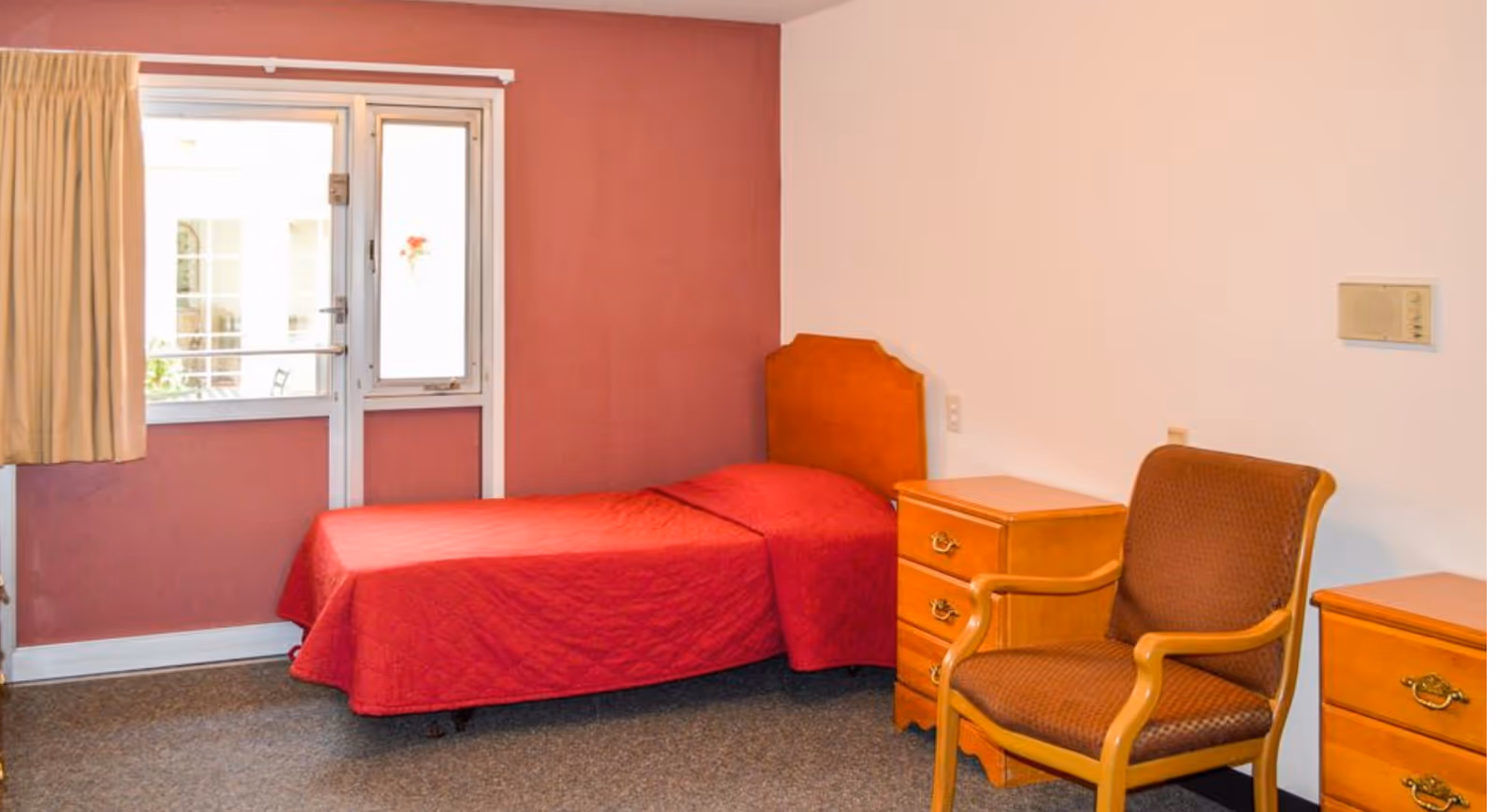 A small furnished bedroom with a red bed, window, and wooden dressers.