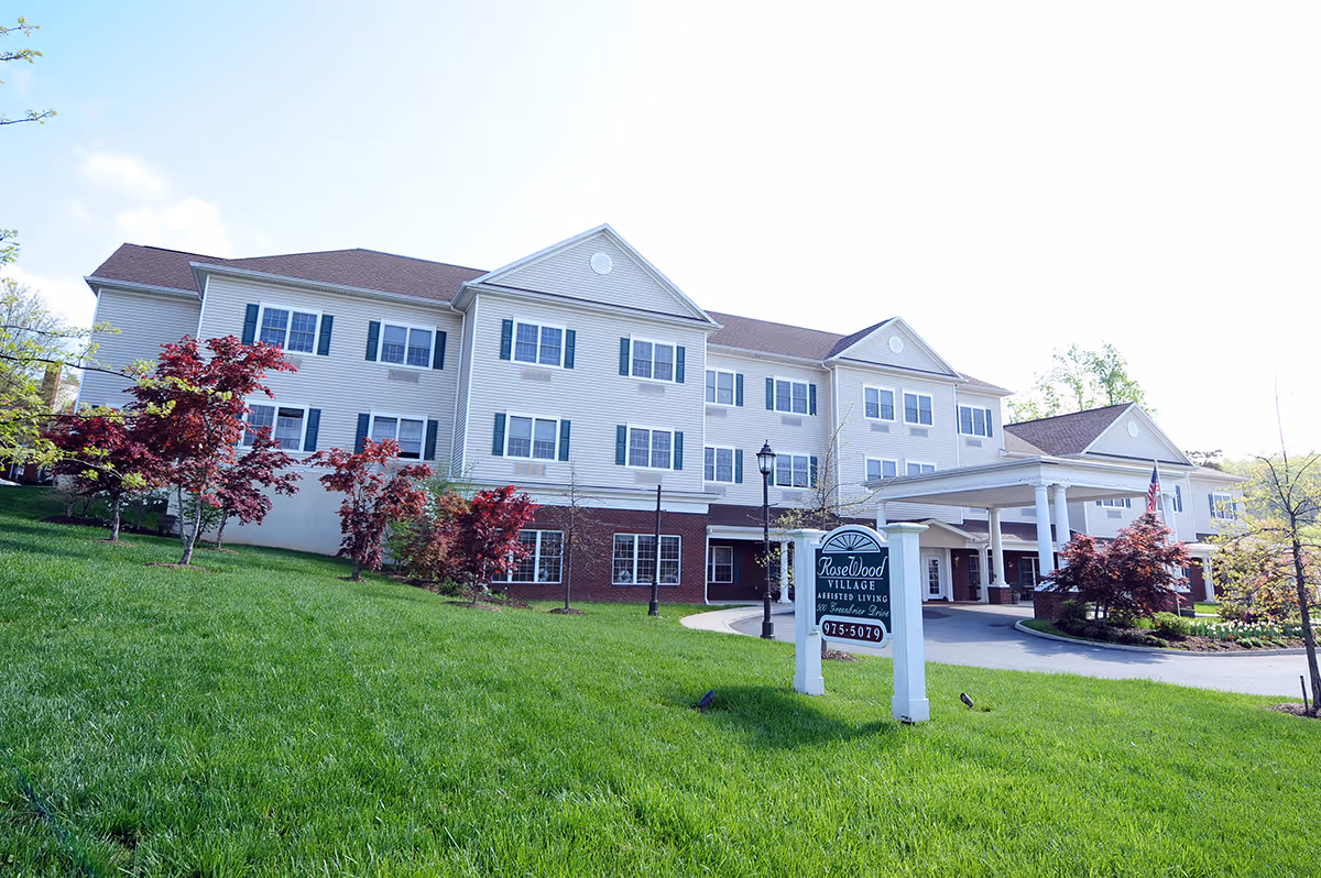 Front exterior of a three-story assisted living building with a covered entrance, landscaped lawn, and a freestanding 'RoseWood Village' sign.