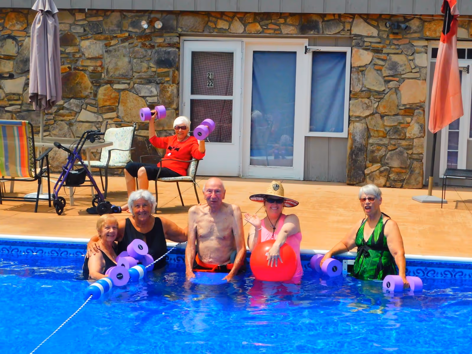 A group of six elderly people enjoying a sunny day at a swimming pool. Four are standing in the pool holding purple foam dumbbells and a red exercise ball, while one person sits on a chair on the pool deck also holding foam dumbbells. The background shows a stone wall with doors and windows.