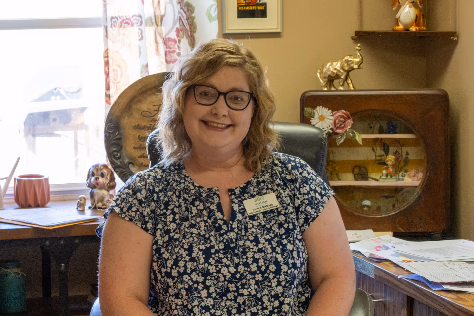 A smiling woman with glasses and curly blonde hair wearing a blue floral blouse and a name tag sitting in an office chair. Behind her is a window with floral curtains, a wooden desk with papers, and decorative items including a small dog figurine and an elephant statue on a shelf.