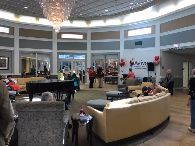 A spacious senior living lobby with curved tan sofas, a grand chandelier, and residents socializing.