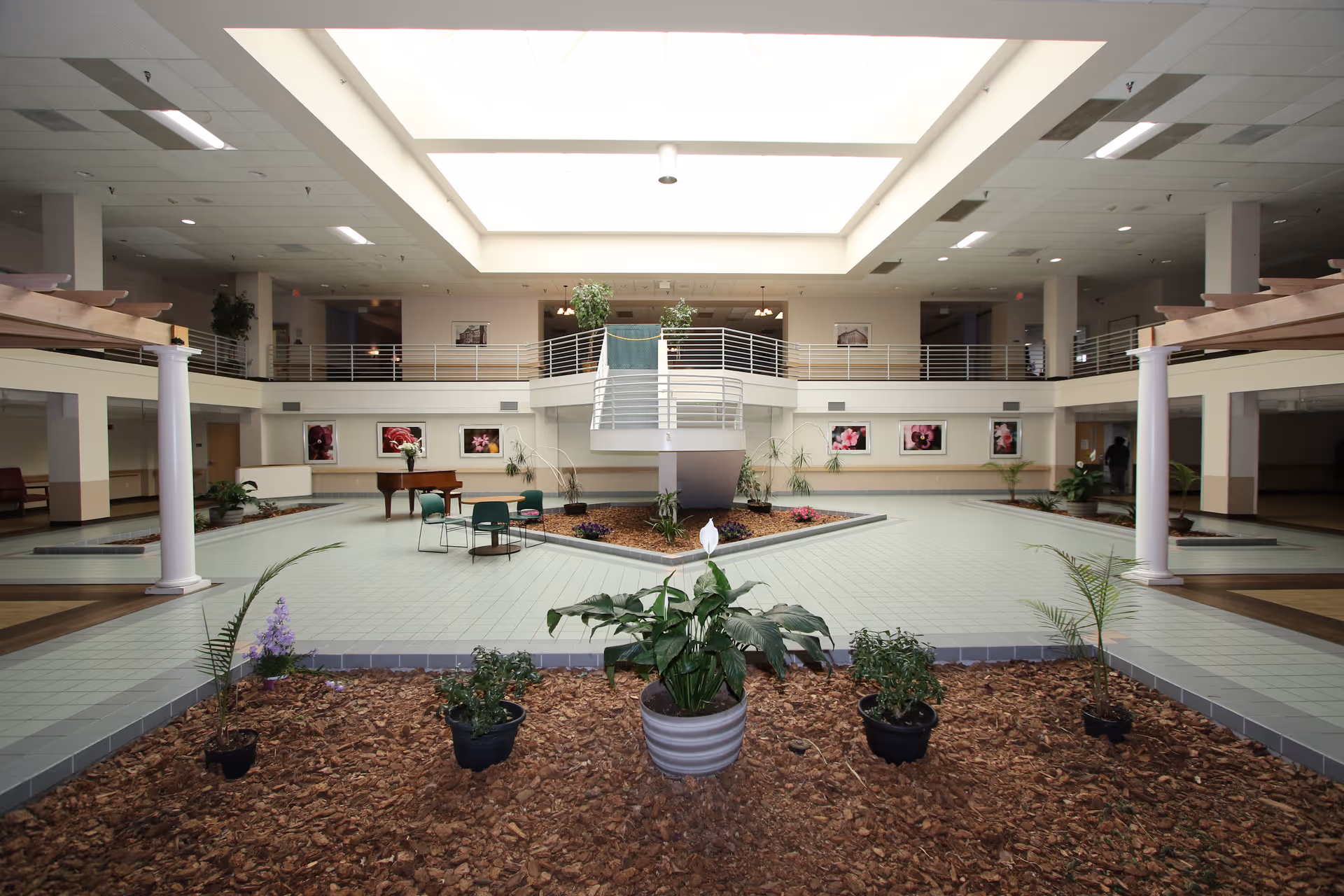 Spacious indoor common area with a large skylight ceiling, surrounded by a second-floor balcony. The floor features tiled walkways and garden beds with plants and mulch. There are chairs and a table near the center, and framed floral pictures on the walls.