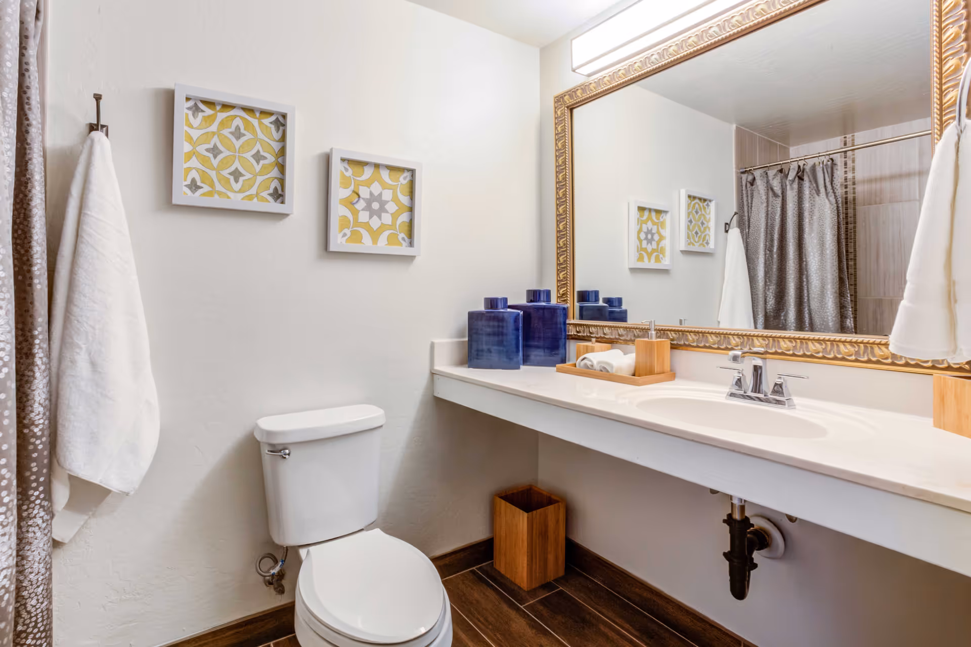 A bright bathroom featuring a toilet, a long vanity with sink and large ornate mirror, blue accessories, and a shower with a curtain.