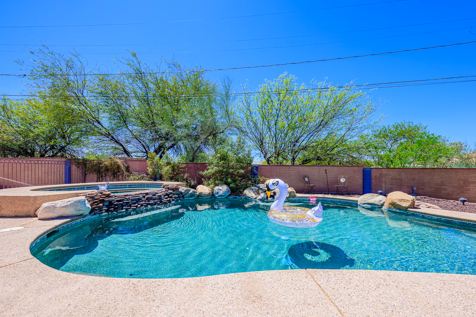 Outdoor pool area with a swan float, bright blue water, stone feature, and trees.