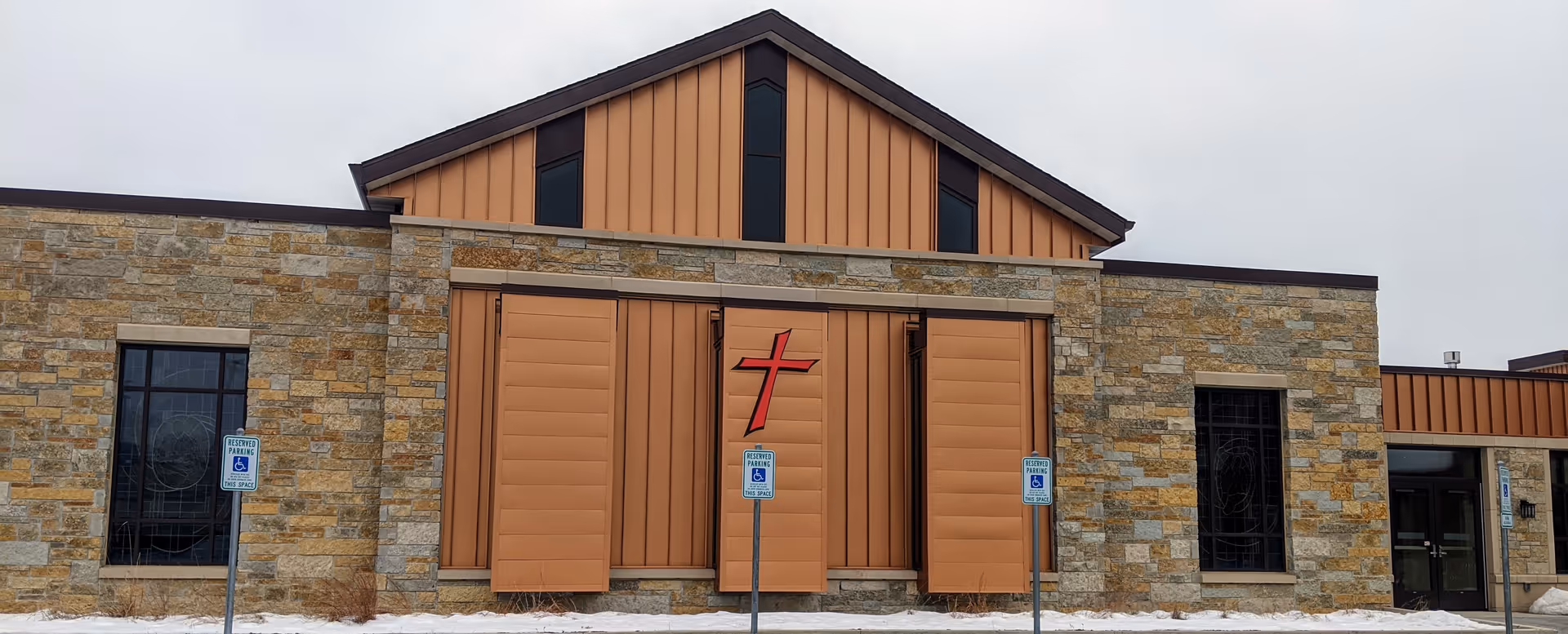 Front exterior of a stone and orange metal-clad building with a cross emblem.