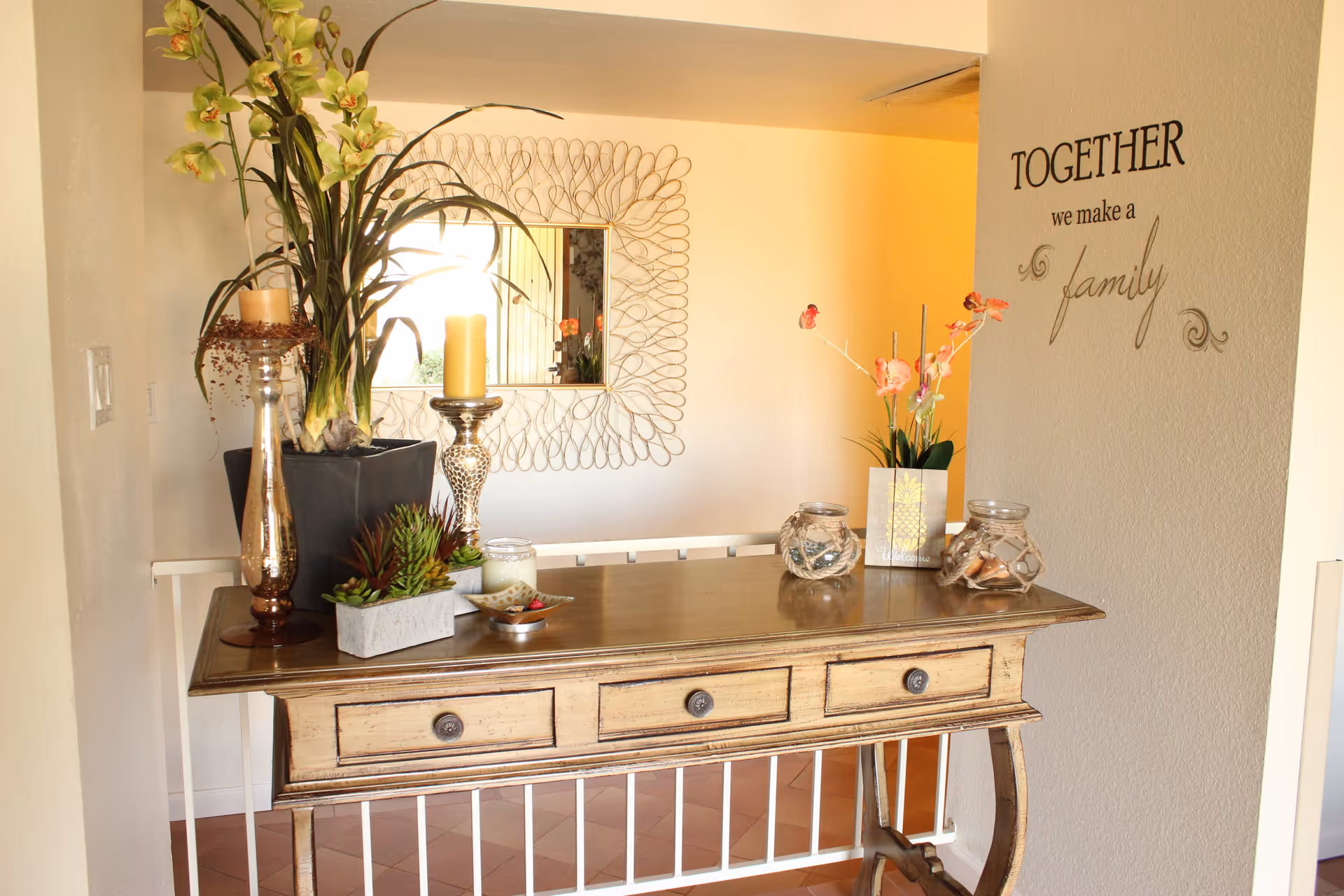 A wooden console table with three drawers is decorated with various items including a tall plant in a black pot, two candles on silver candle holders, small succulent plants, a small dish with decorative items, two glass jars with rope accents, and a small floral arrangement in a vase. Behind the table, there is a decorative mirror with a unique looped frame hanging on the wall. On the adjacent wall, a decal reads 'TOGETHER we make a family'. The setting appears to be a warmly lit interior space.